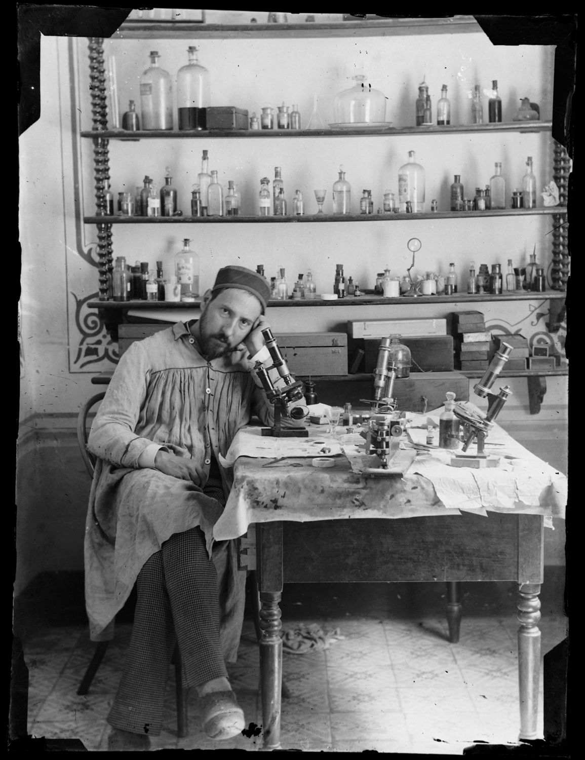 Santiago Ramón y Cajal sitting by microscope with various bottles on shelves behind him