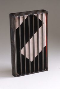 A book resembling the Nazi flag, with thick black and white rectangular lines, and a hint of red, is enclosed in a metal cage.