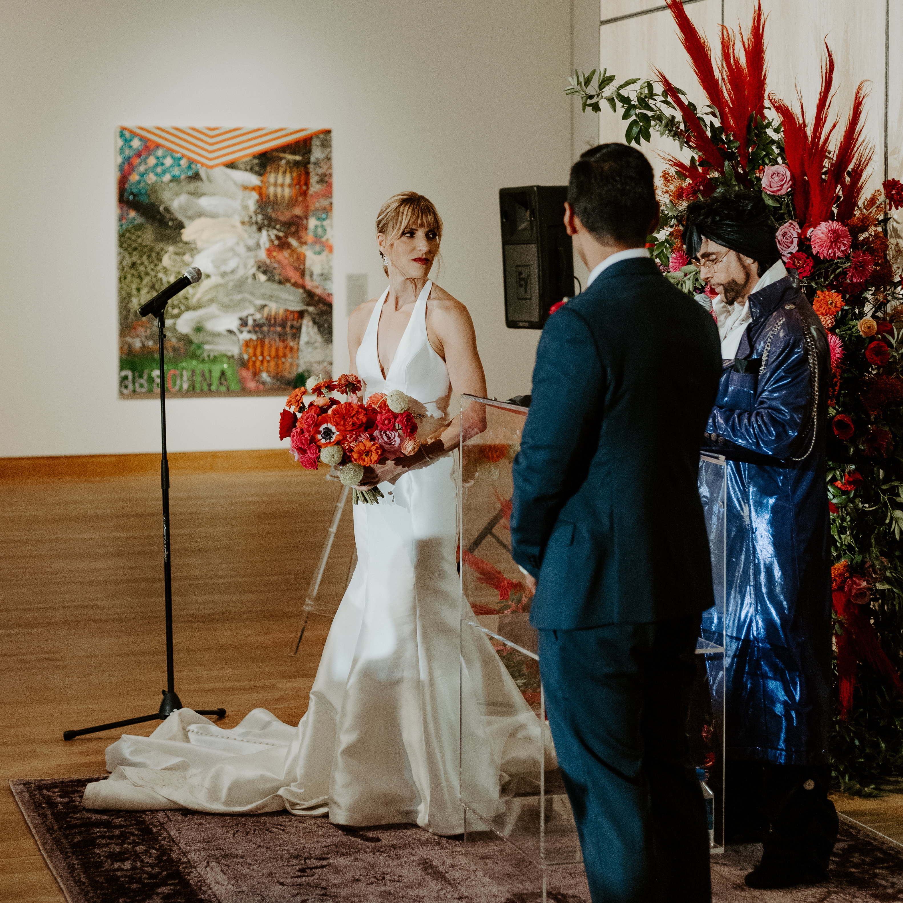 A femme stands in a white bridal gown, in the middle of an art gallery, holding a bouquet of red flowers. A masculine figure in a dark blue suit stands with his back to the camera. A Prince impersonator stands between them in a sparkling purple suit. 