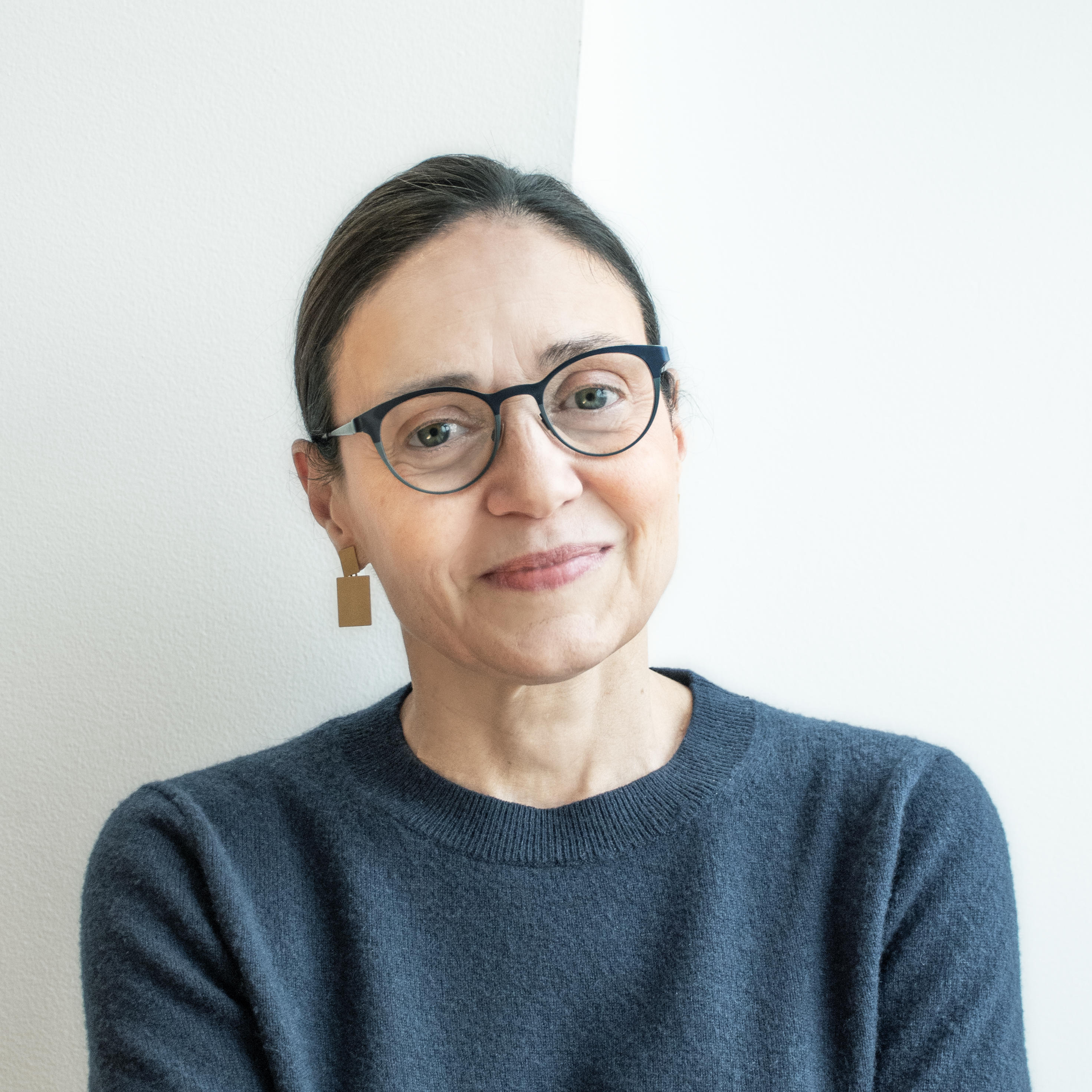 A older light skinned femme with dark brown, shoulder-length hair smiles circumspectly at the camera. Diane is wearing dark blue glasses, large golden and minimal rectangular earrings, and a navy blue crew-neck sweater. 