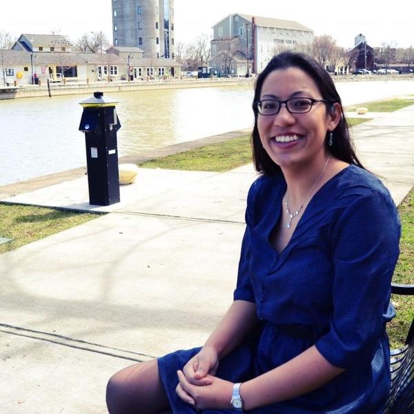 With long dark hair, wearing a long sleeve blue dress and glasses, Mariam Banahi, the author, is seated on an iron bench outside by a waterway; she's facing the camera with a big smile.