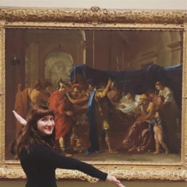 Portrait photo of Laura with her arms open in front of a framed painting at a museum.