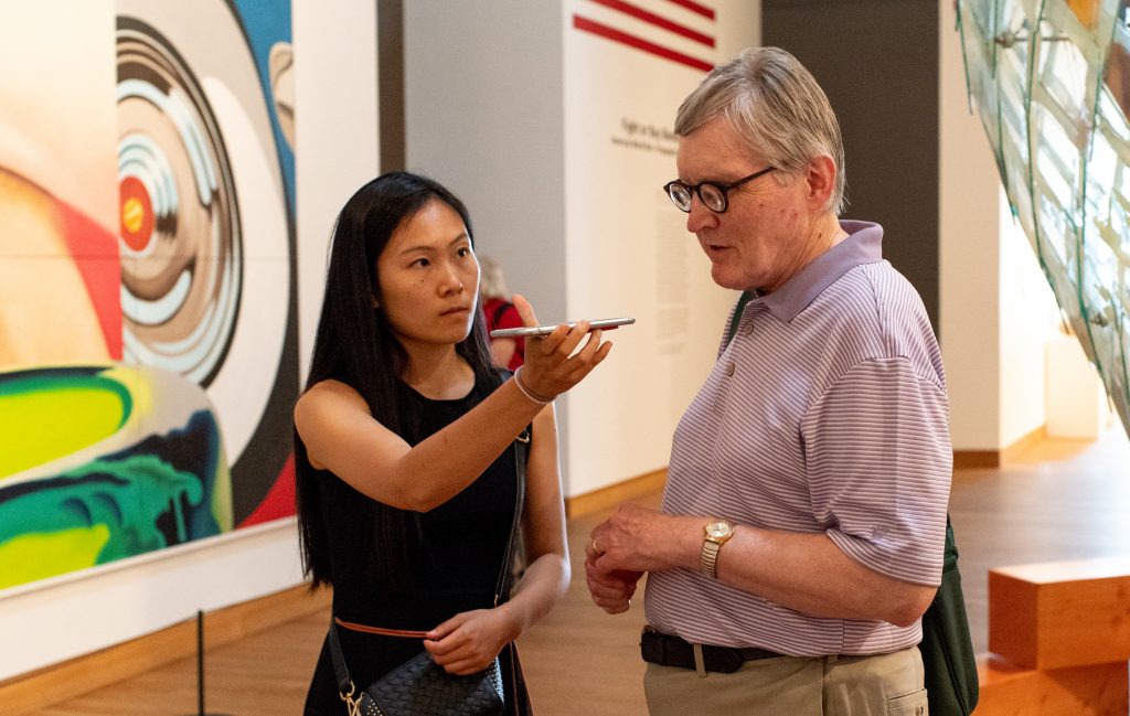 Two people are in an art gallery, a woman and a man. The woman holds a phone up, in front of the man. Both are exploring accessibility resources as part of "The Sensory Loss Symposium" at the Weisman in 2018.
