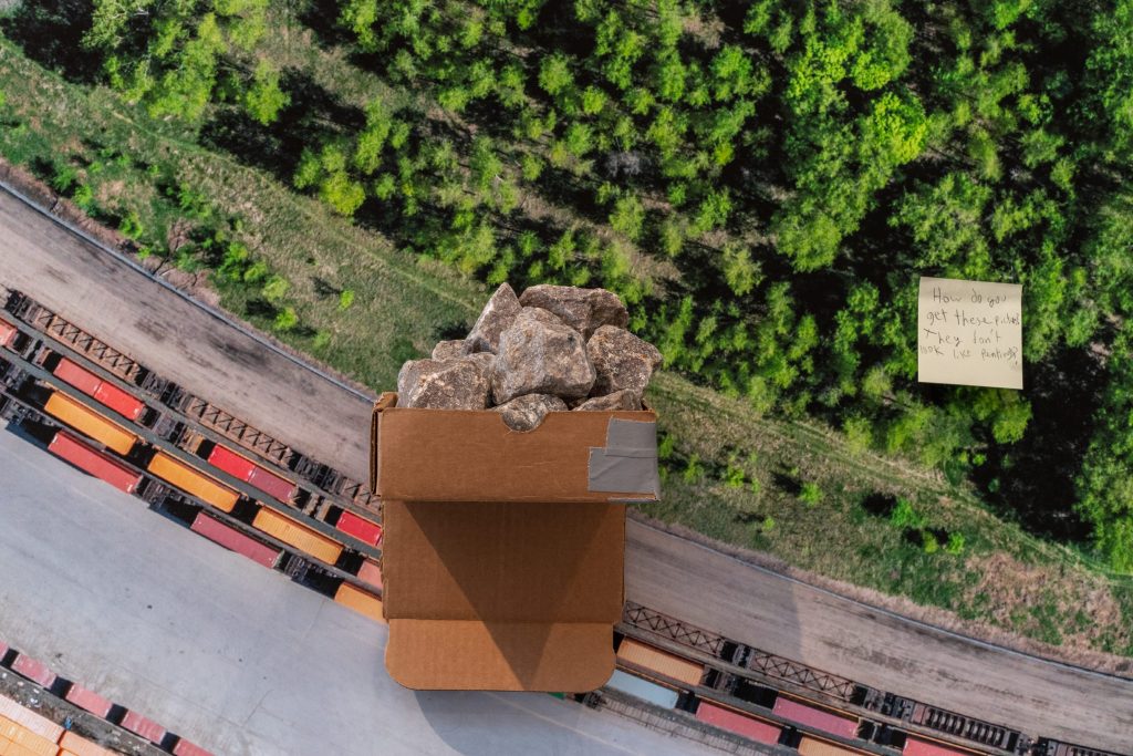 Detail of exhibition collage, with cardboard box filled with gray rocks and an aerial view of the trainyard and marginal forest