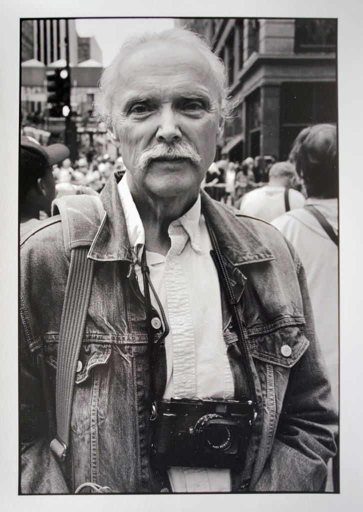 A lanky man with a bushy white mustache and swept-back white hair faces the viewer, camera around his neck. He's wearing a denim jacket and crisp white button-down shirt and is pictured standing, from the waist up, on a crowded city street.