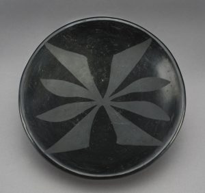 Black sheen plate with matte black geometric and symmetric design in the center and extending outwards, covering the surface of the plate.