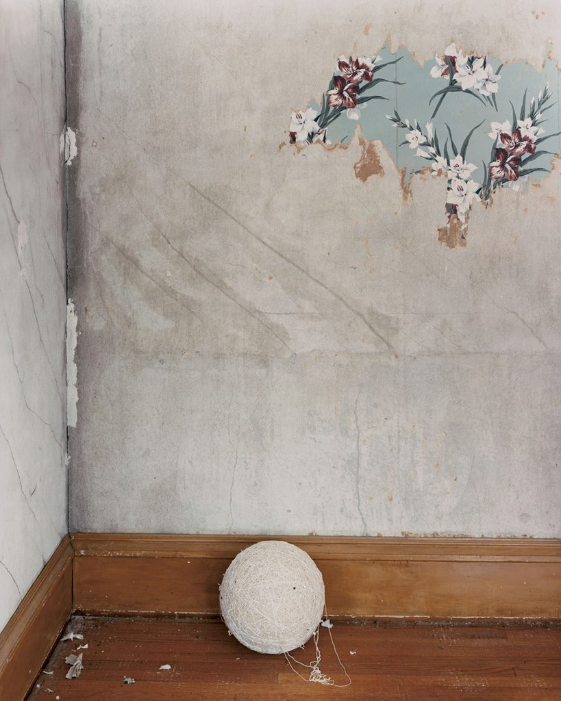 A photograph of a ball of string on a wooden floor in the corner of a room; a bit of floral wallpaper remains on an otherwise blank wall.