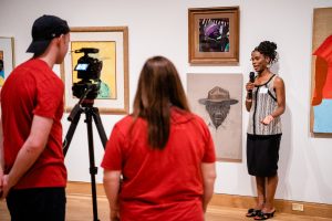Artist Maya Beecham—a family member of Rose Smith— stands in front of Rose's paintings and provides an interview to two StoryMobile team members who record her using a camera on a tripod.