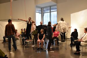 Dancers of all ages who wear hospital gowns with plastic gloves attatched are watched by a seated audience in the Target Studio Gallery at the Weisman.