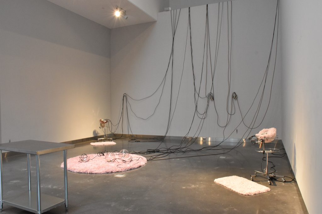 View of installation, As Seen, with pink fuzzy rugs, and black wires lining the wall. 