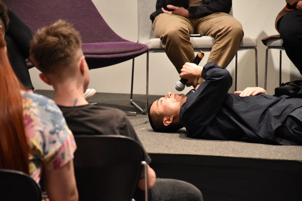 Peng Wu lies on his back on stage, looking up to the ceiling; the frame includes two audience members watching him.