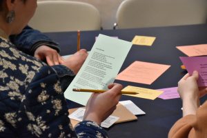 A workshop participant picks up a piece of paper with a poem on it and a pencil. The table is filled with colorful pieces of paper.