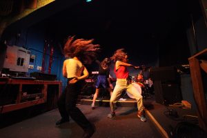 Three DaNCEBUMS movers dancing in the center of a stage, swinging their arms and hair.