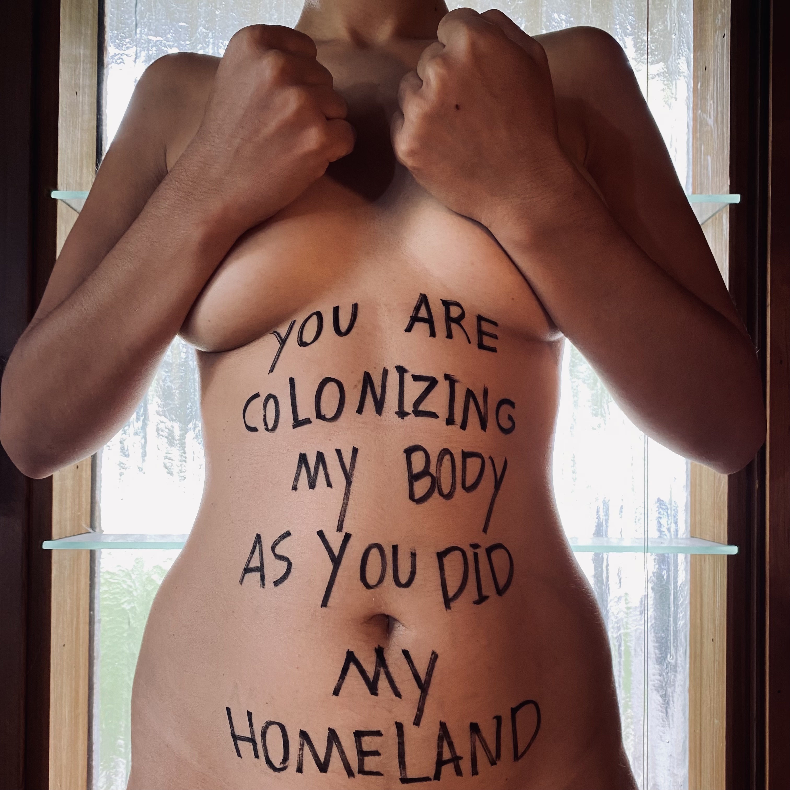 A naked, female-presenting torso faces the camera in the foreground, in front of a window; her arms partially cover her breasts; her fists are clenched and poised up, as if ready to punch. On her belly is a message written in thick, black, all-caps: YOU ARE COLONIZING MY BODY AS YOU DID MY HOMELAND