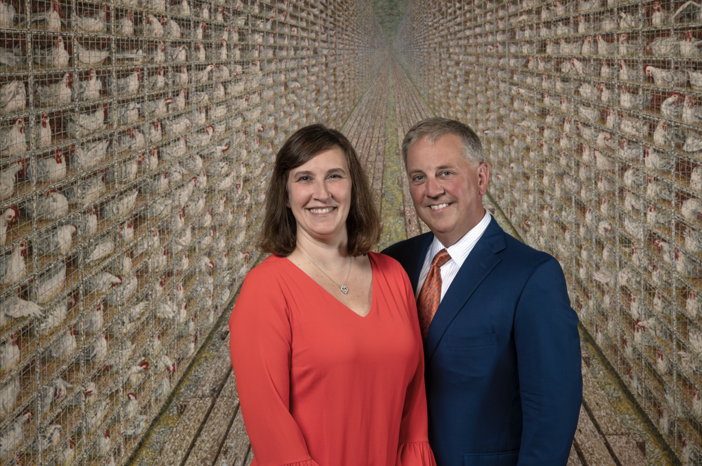 2 people standing in front of rows of chicken cages