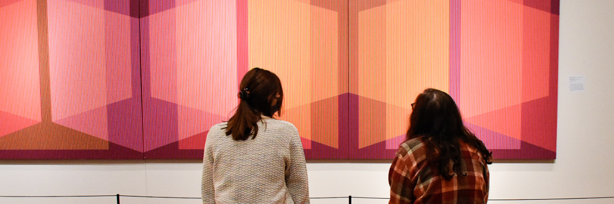 two people observe large art piece on wall