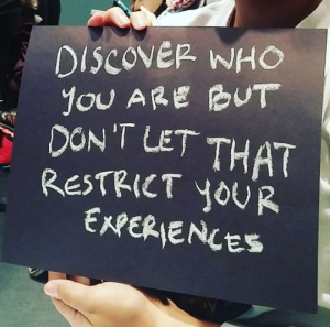 A person holding a sign saying 'discover who you are but don't let that restrict your experiences'