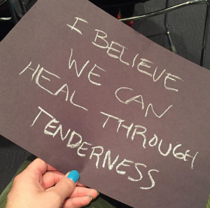 a hand holding a sign saying 'I believe we can heal through tenderness'