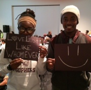 2 people holding signs, one saying 'love lust like whomever' and the other with a smiley face