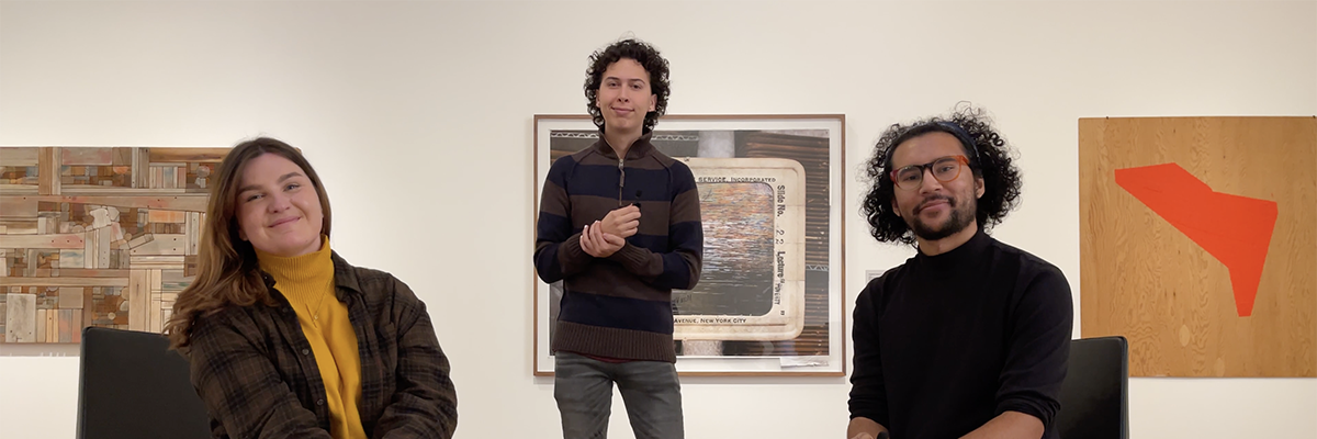 Three people in an art gallery look at the camera. The people in the photo look like: a long-brown haired femme wearing a yellow sweater and flannel jacket, a thin non-binary person with long, dark curly hair and light skin, a medium-brown skinned non-binary person with medium length black curly hair and a beard. 