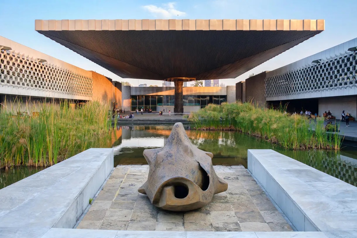 A modern looking courtyard with a large shell sculpture on a platform of marble and stone. There are tall green grasses surrounding the sculpture which emerge from a rectangular pond.