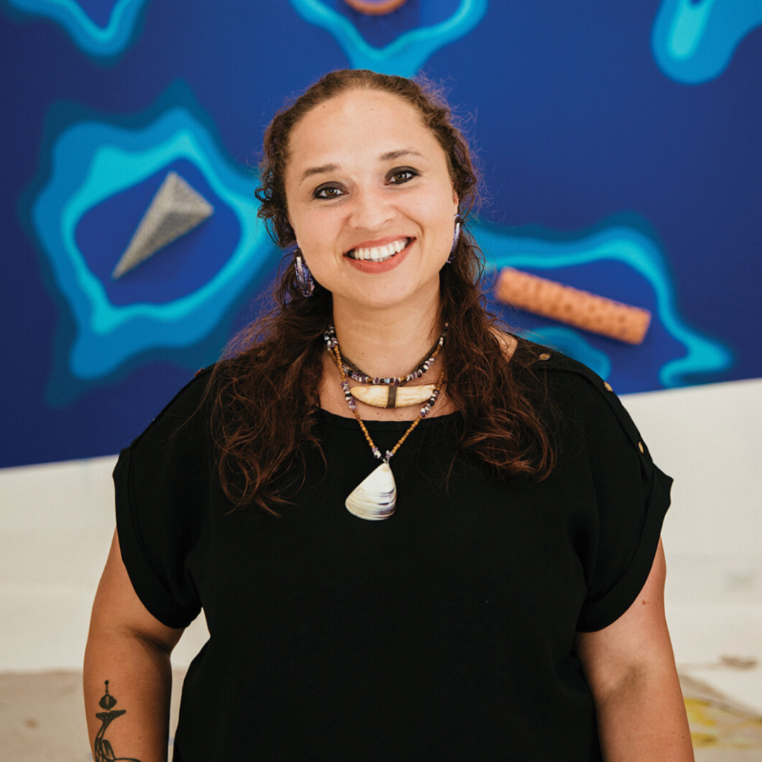 Light-skinned, femme with long, curly brown hair stands in front of a blue-painted wall with ceramic baskets and nets placed on radiating "puddles" of blue paint. 