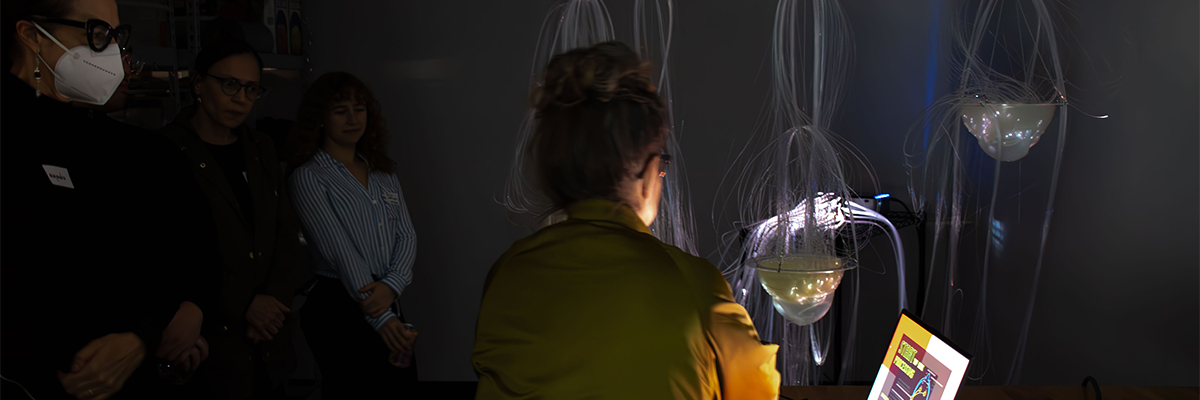 A woman with dark hair and a green shiny jacket shows a small group of people her artwork in a darkened room. There are fiberoptic tubes spilling out of a glass dome (illuminated by the artist's open computer screen). 
