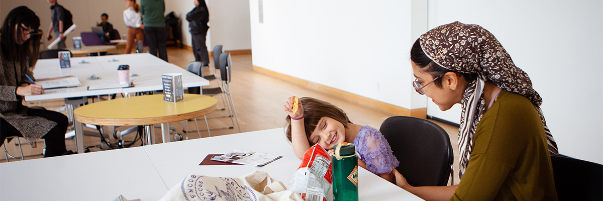 A child eating snacks, joyfully, with their femme parent in the Riverview Gallery. 