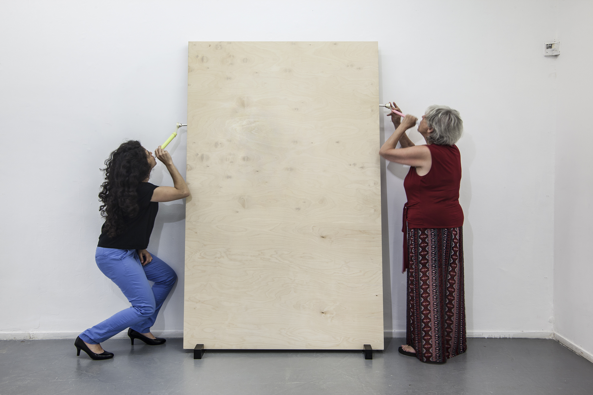 Two femme presenting people appear to blow into a slab of concrete through straws. 