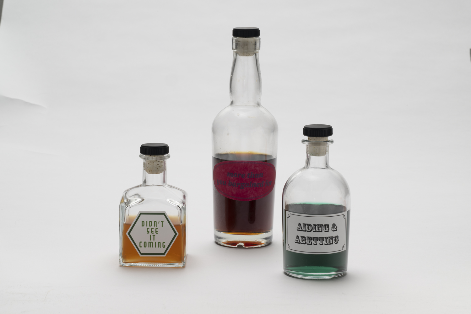A trio of bottles, each a different size and shape, are partially filled with liquids: one amber, one deep red, one mint green. The bottles are each labeled according the scent of the liquid they contain: "Didn't See it Coming," "More than you Bargained For," and "Aiding & Abetting"