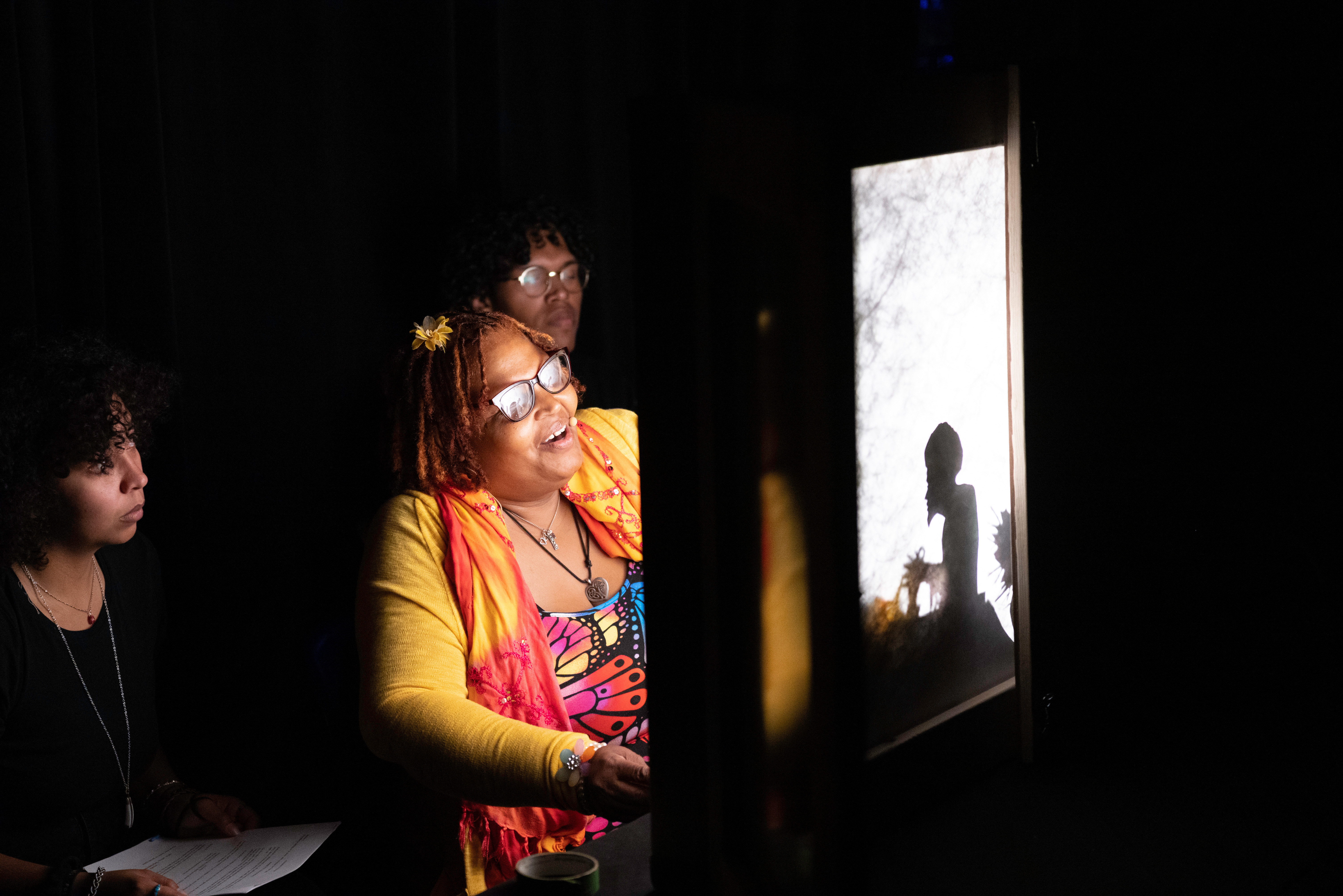 The faces of three dark-skinned, Black individuals are lit up by a light box with a shadow puppet inside of it. The central figure is a femme with brightly colored clothing (orange and red) while the other two are wearing black. The figures are in a near pitch black room (no other elements are distinguishable). 