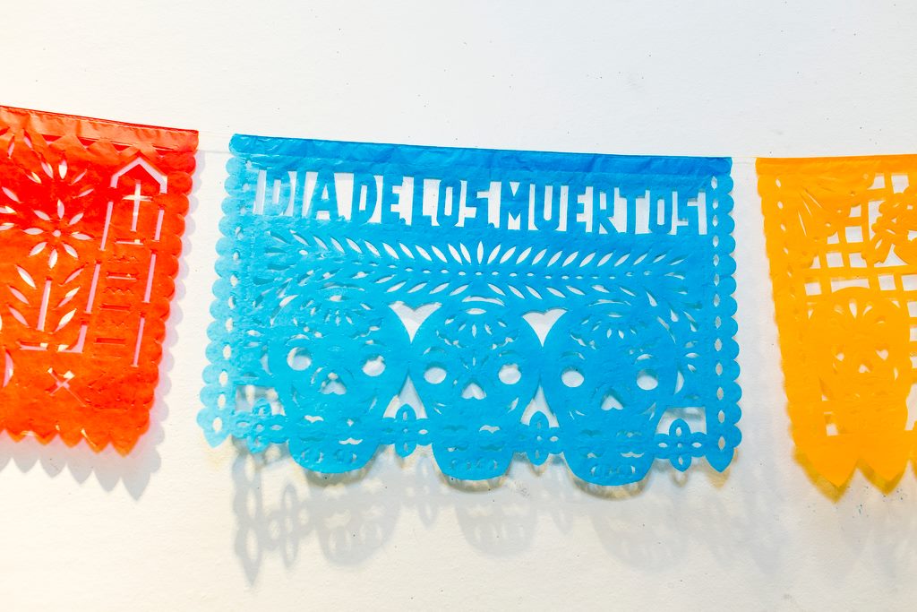 Paper-cut, colorful flags in blue, yellow, and read, with cut-outs reading Dia De Los Muertos