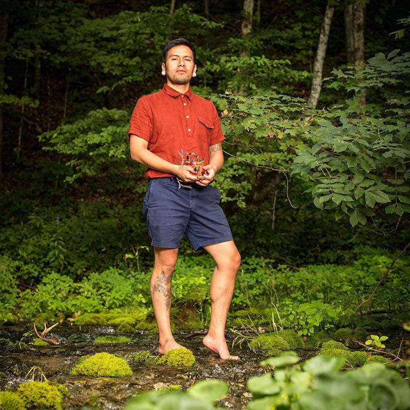 A man with a red t-shirt and dark blue shorts stands, barefoot, in a lush jungle environment.