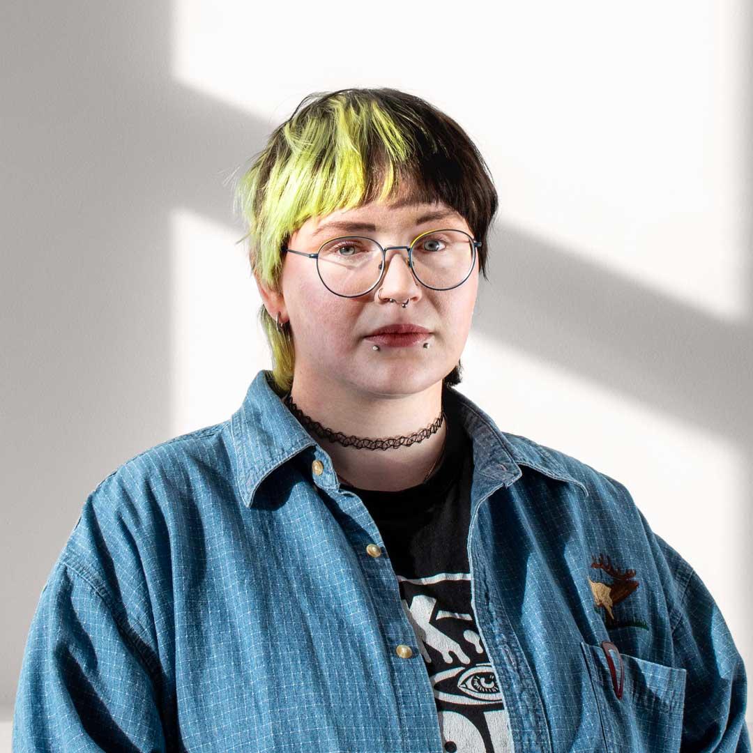 A person with short, dyed electric green and black hair wearing glasses, a black lace choker necklace, a graphic tshirt and a blue denim overshirt looks stoically at the camera. Emma has snakebite, septum, and nose-ring piercings.