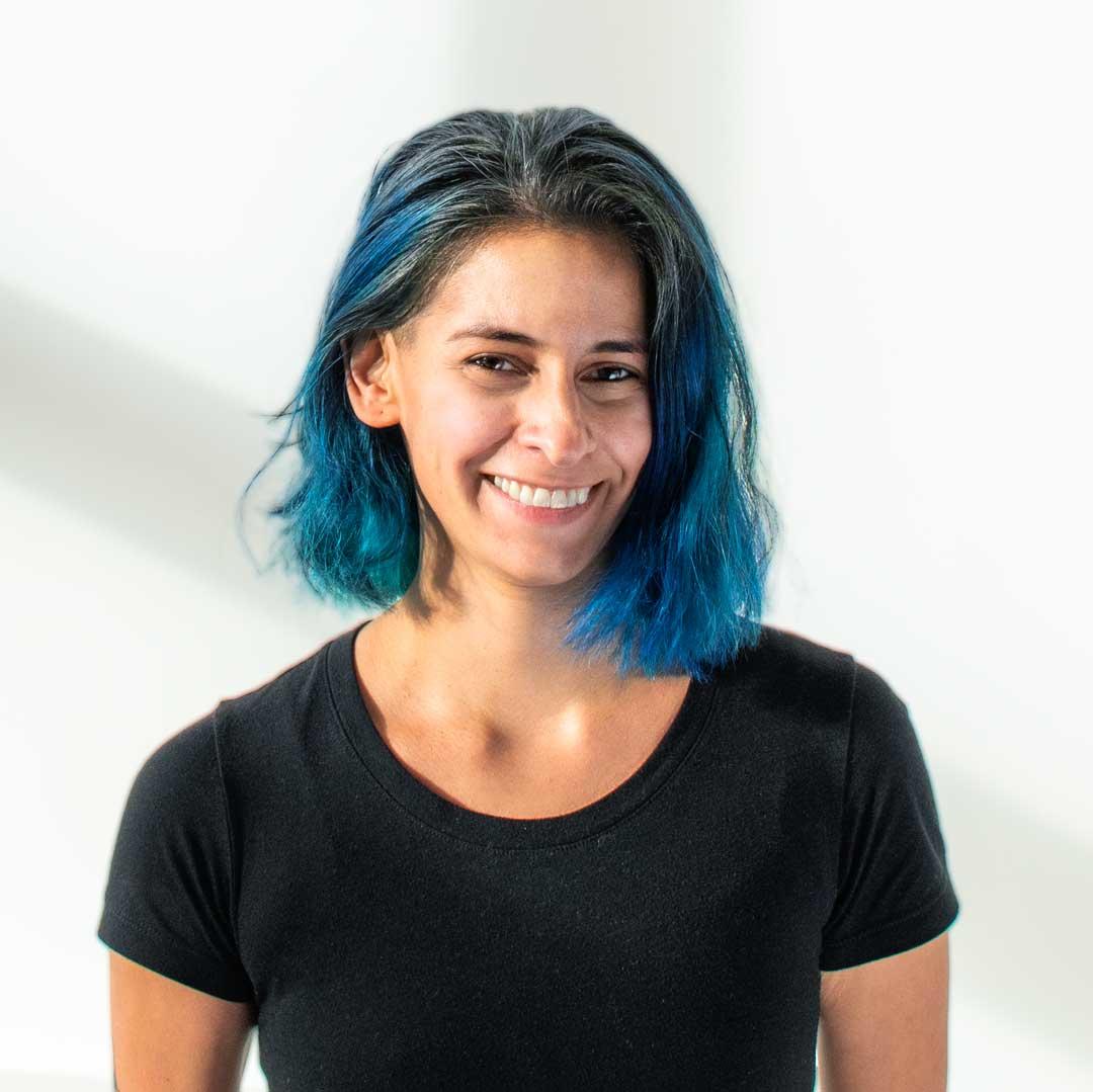 A femme-presenting person with electric blue hair, light brown skin, and a black tshirt smiles broadly into the camera.