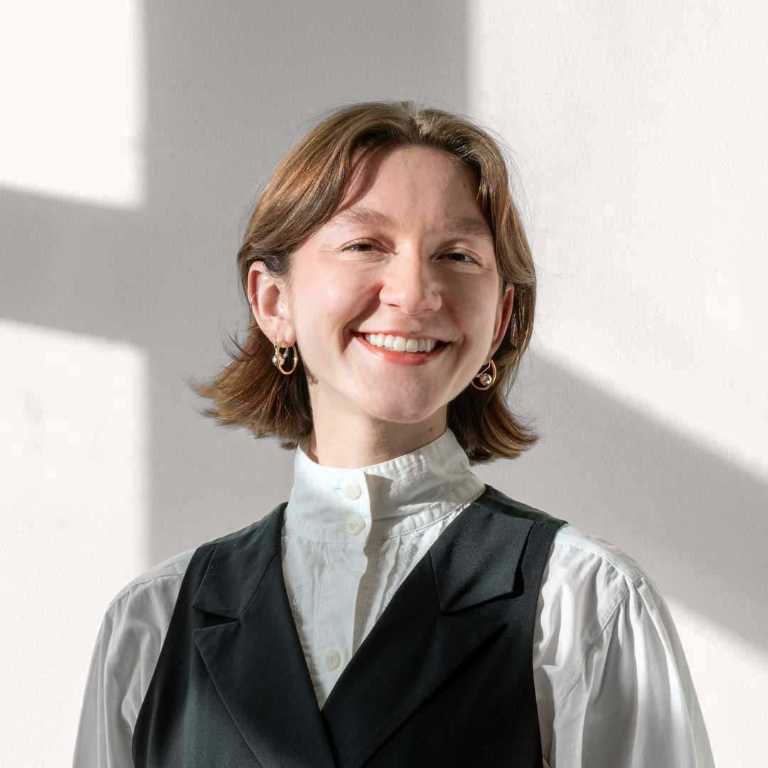 A femme with a brown long bob smiles broadly at the camera. Kate has light skin and medium brown hair and eyes. They wear a white button down shirt and a black waistcoat.