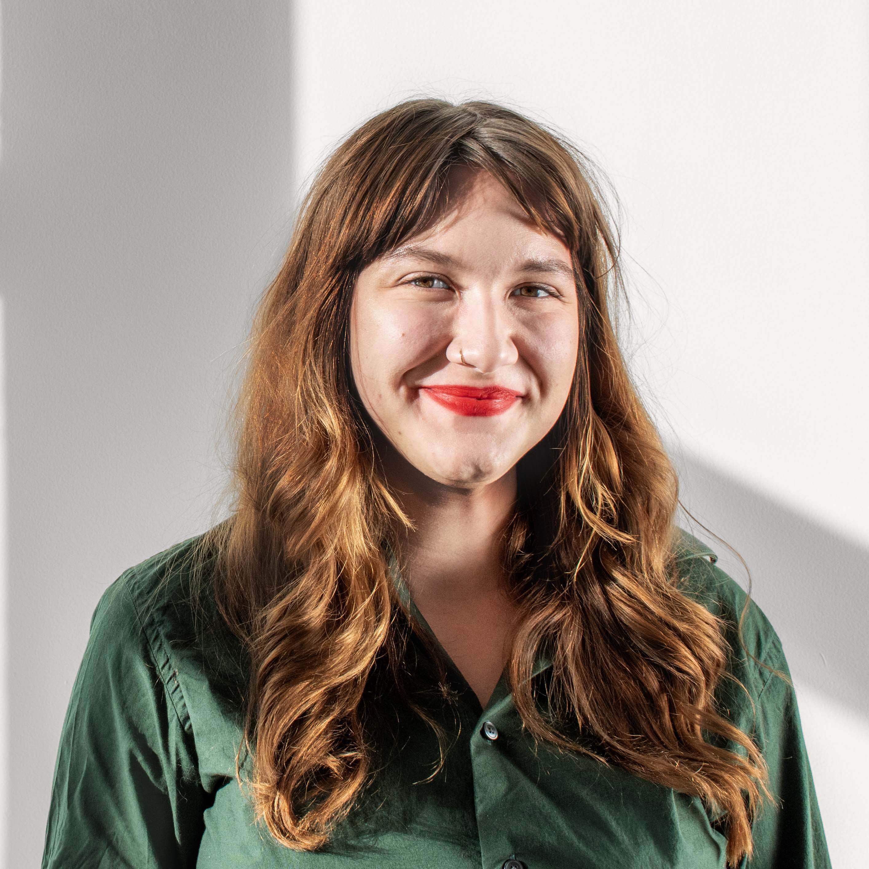 A femme with long, curly, auburn hair and light skin smiles at the camera. Laura wears red lipstick and a green button down shirt.