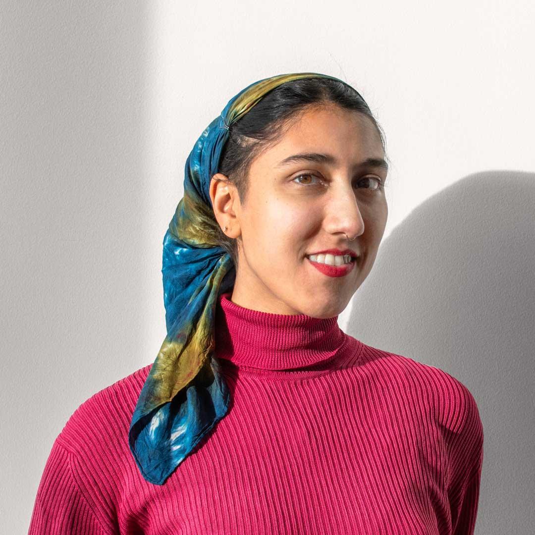 A femme presenting person with light brown skin wears a green and blue tie-dye scarf to cover their hair, a bright pink turtleneck shirt, and red lipstick. Madeeha smiles softly into the camera. 
