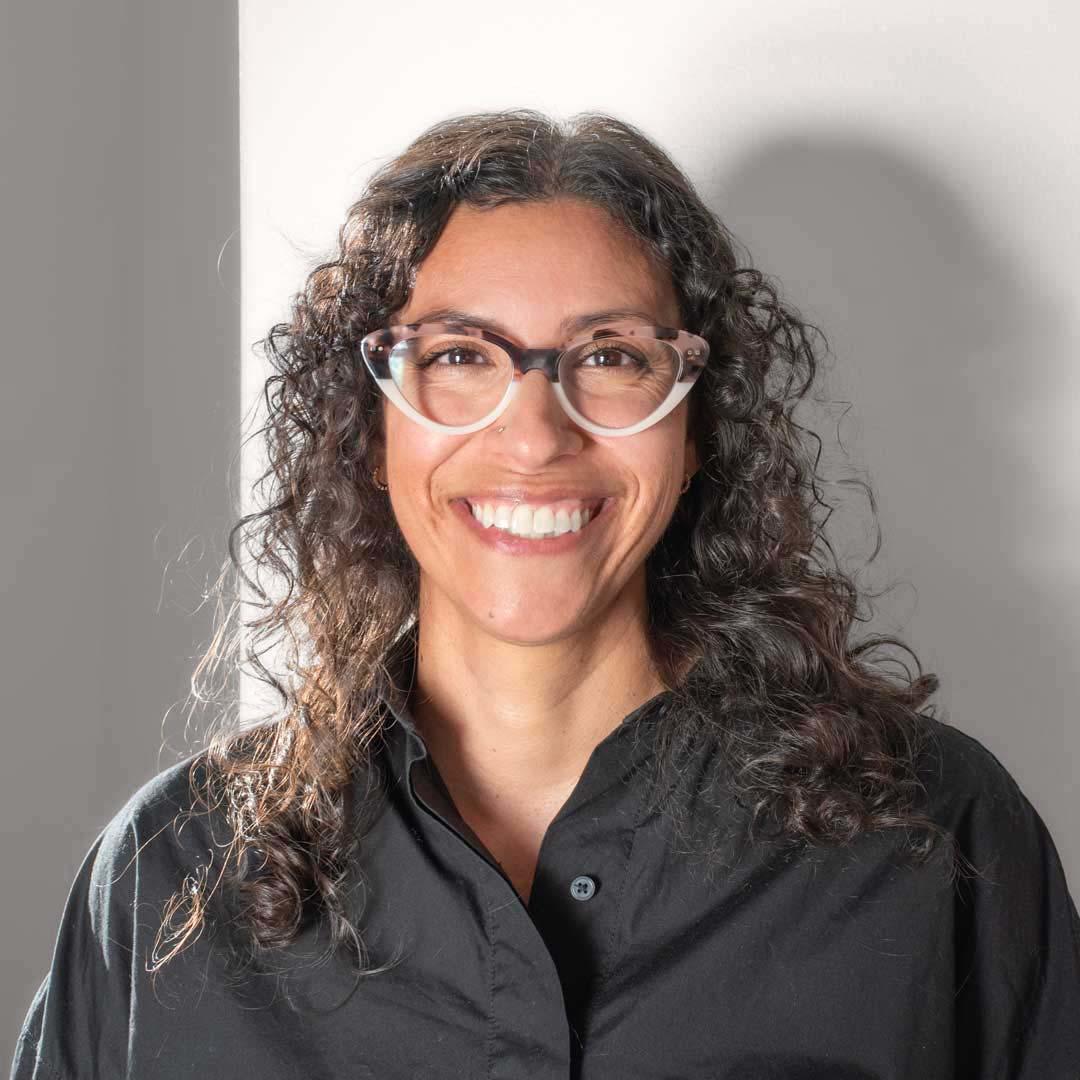 A light brown skin femme smile broadly into the camera. She has curly dark brown hair and half-rimmed tortoise shell glasses and wears a black button up shirt.