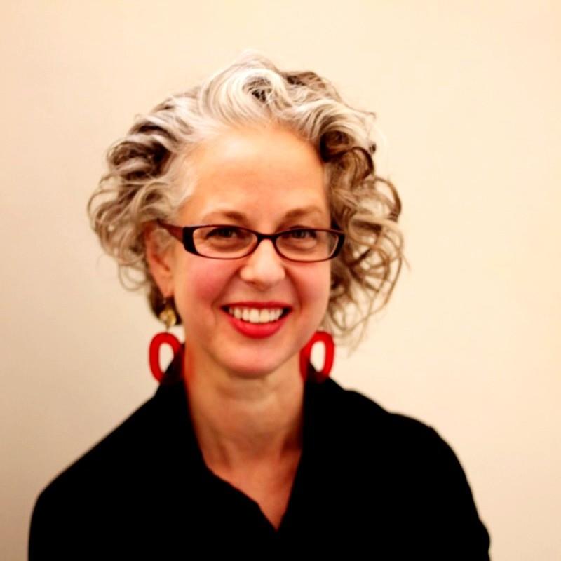 A woman with short curly hair, a black shirt, and glasses smiles into the camera