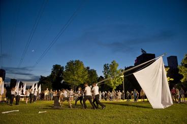 A nighttime color photo depicts four people in white shirts and black pants raise a large white flag,surrunded by spectators at Northern Spark festival in Minneapolis. 