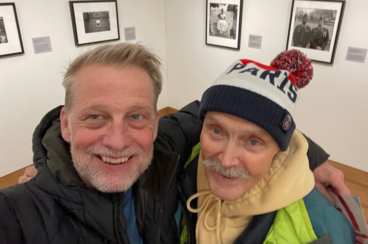 Two men are pictured in a gallery space, their arms thrown over each other's shoulders, smiling for the camera. They are dressed in winter coats; the man on the right has a stocking hat with the word "Paris" emblazoned on it.