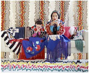 Wendy Red Star and her daughter are seated on a couch covered in blanket, quilts, and other textiles. A colorful, patterned background is behind them, with a different colorful, patterned background on the floor.