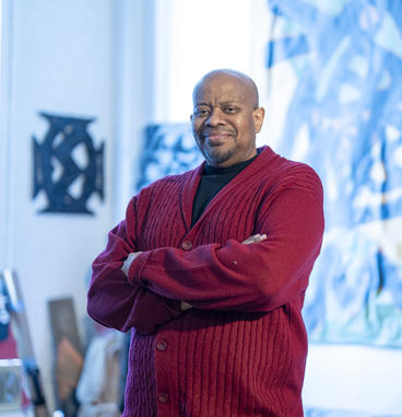 A man with very short dark hair and a trimmed beard, wearing a red jacket, pictured in an art studio. 