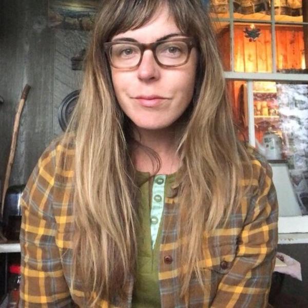 Shanai looks straight at the camera. She has long brown hair and glasses and is wearing a flannel shirt layered over a light green knit shirt.