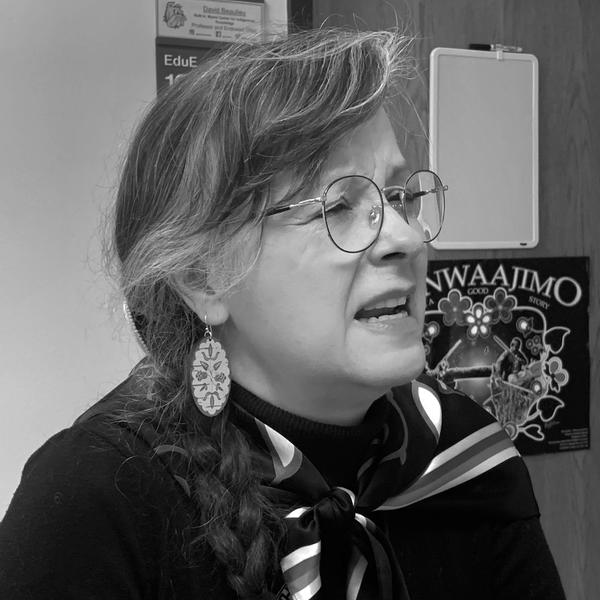Black and white image of a light-skinned femme with graying hair and round eyeglasses. She looks off camera and appears to be speaking.