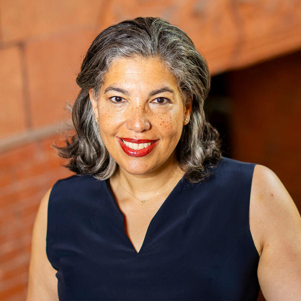 A light-skinned, Black femme with salt and pepper, shoulder length hair smiles with teeth into the camera. Gwendolyn DuBois Shaw wears red lipstick and a dark, navy blue dress.