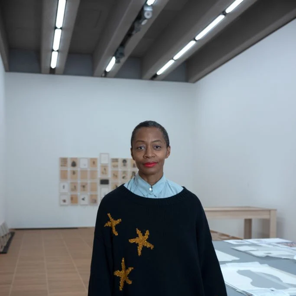 A Black woman with hair in a tight bun and red lipstick, looks into the camera with a penetrating gaze. She wears a light blue shirt, an oversized black sweater with a few large gold stars knitted into it and floral pants. Kara Walker stands in a gallery with her prints on a table out of focus behind her. 