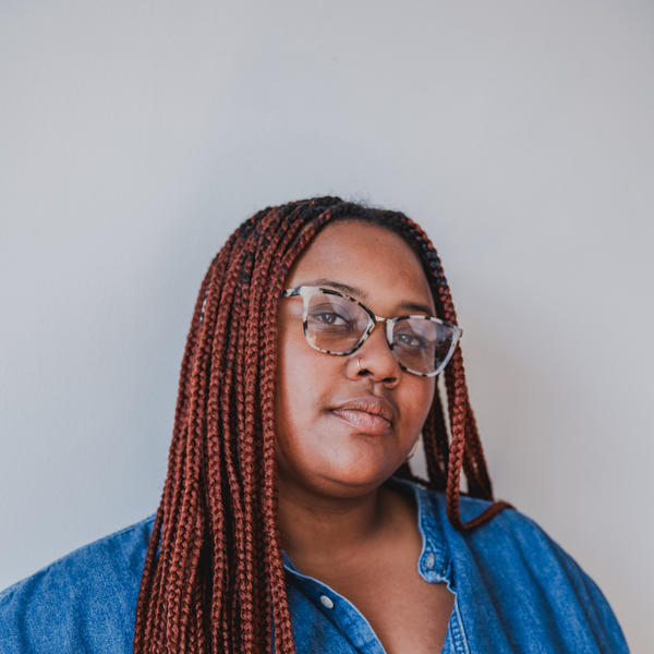 A femme-presenting person with long locs and glasses, wearing a blue button-up, looks into the camera