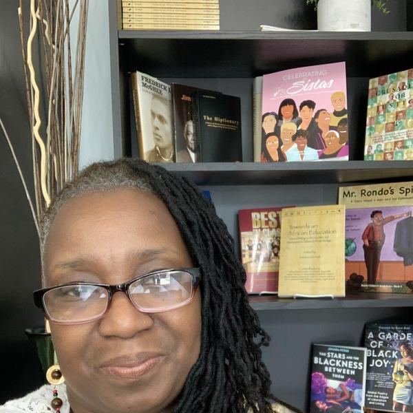 A woman with long locs, pulled over one shoulder, and wearing glasses sits in front of a bookcase